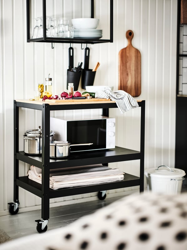 Carrello BROR in nero/compensato di pino con forno a microonde TILLREDA bianco, sotto una struttura da parete con ripiani ENHET antracite. Sul piano di lavoro ci sono diversi alimenti e due contenitori SKATTÅN. Sopra i contenitori uno scaffale ENHET sui cui ripiani ci sono bicchieri e ciotole OFTAST. Accanto al carrello un tagliere ARTISTISK appeso al muro e un bidone con coperchio KNODD.