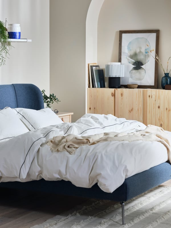 Stilrent sovrum med blå säng, vit bäddning och beige filt; träskåp med dekorationer och hylla med växter i bakgrunden.