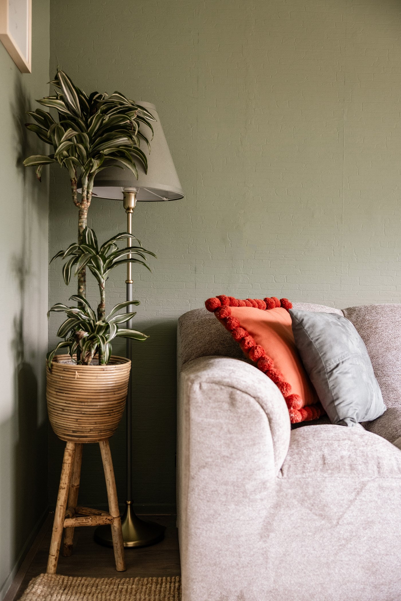 Close-up van een woonkamer met grijze bank, en daarnaast een lamp en houten krukje met daarop een plant.