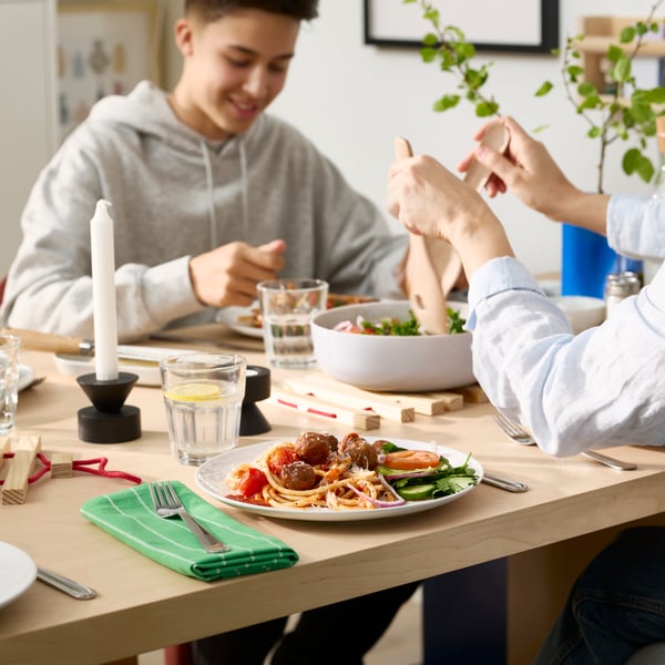 Two people are sitting at a dining table and eating food from white GODMIDDAG plates and drinking water from POKAL glasses.