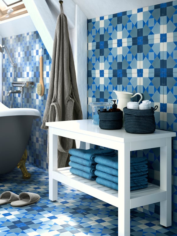 A white HEMNES bench with towels on a lower shelf and baskets with accessories on top, with blue tiled walls and floor.
