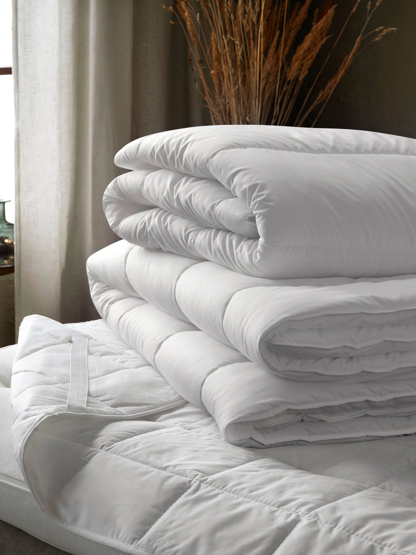 Two white, folded duvets in a pile on top of a bed with a mattress protector. A window with light curtains in background.