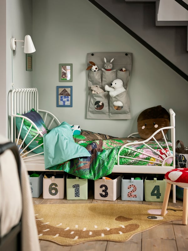 Lit d'enfant extensible MINNEN dans un coin de la pièce, avec des boîtes de rangement numérotées en dessous et des peluches au-dessus.