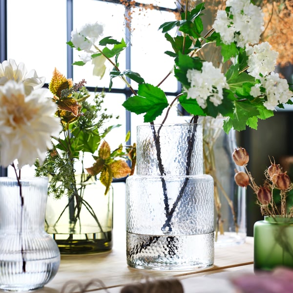 A green-brown KONSTFULL vase and a KONSTFULL transparent glass vase with flowers next to a window. 