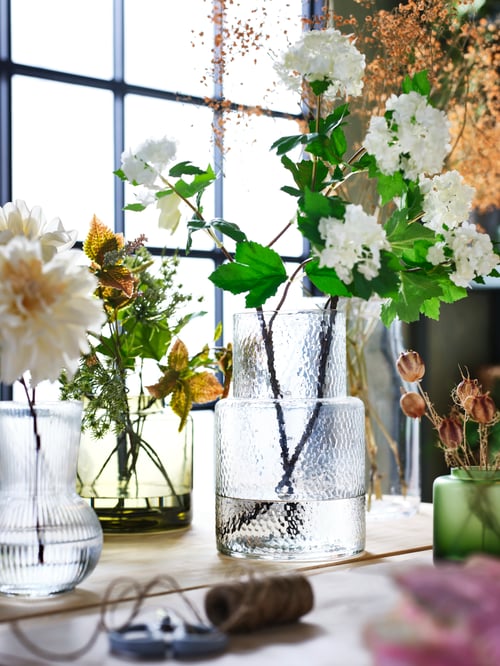 A living room featuring a clear glass KONSTFULL vase, a green-brown vase and a PÅDRAG vase, all of them filled with flowers.