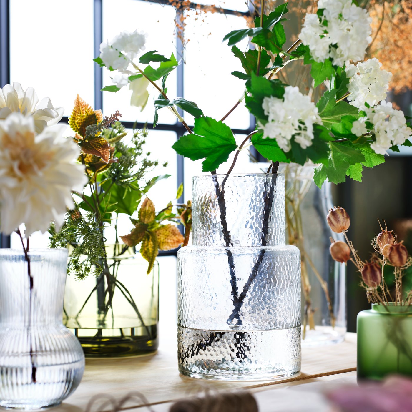 A green-brown KONSTFULL vase and a KONSTFULL transparent glass vase with flowers next to a window. 