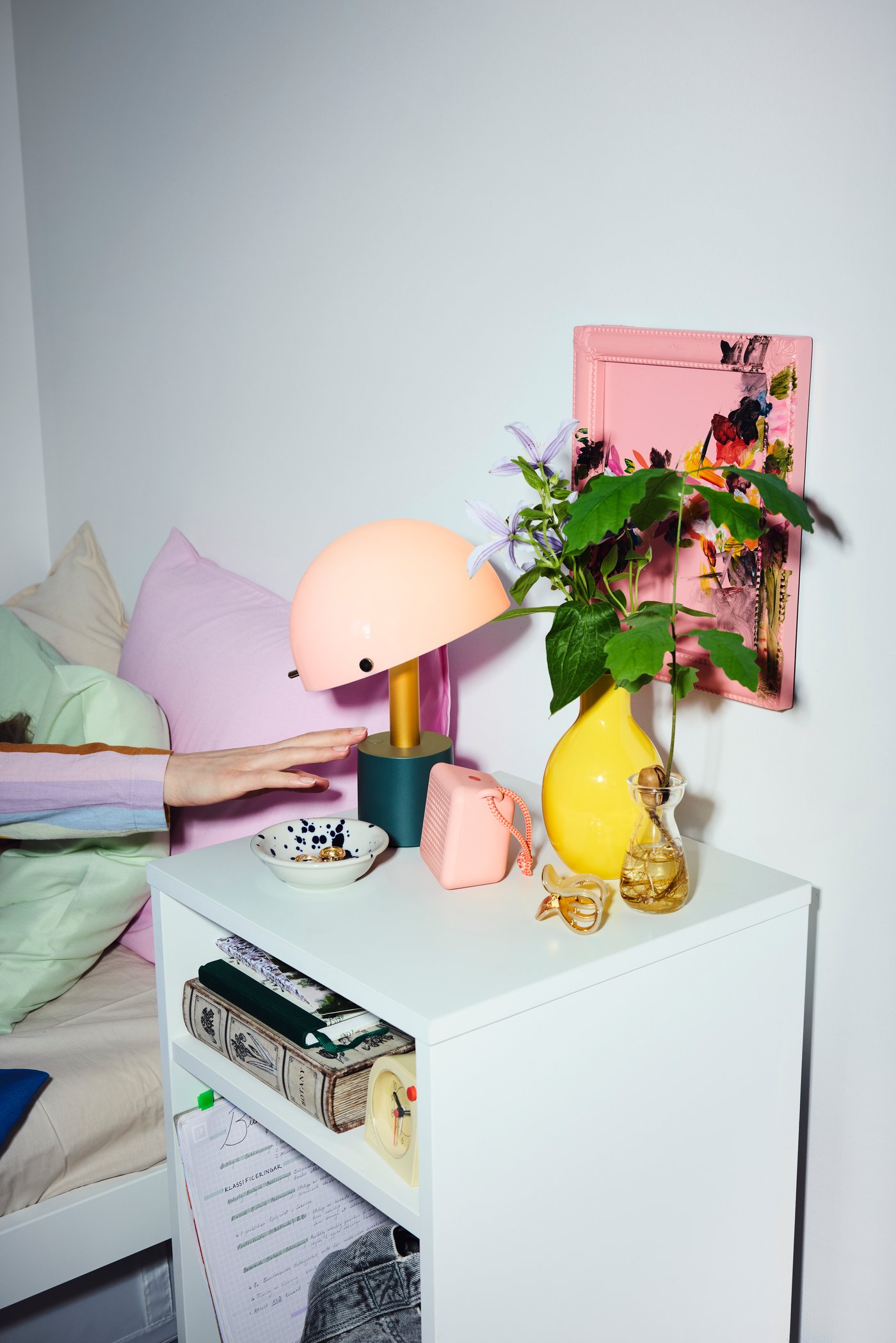 En hånd rækker ud efter en lyserød og mørkegrågrøn NÖDMAST bærbar LED lampe, der står på et sidebord i et soveværelse.