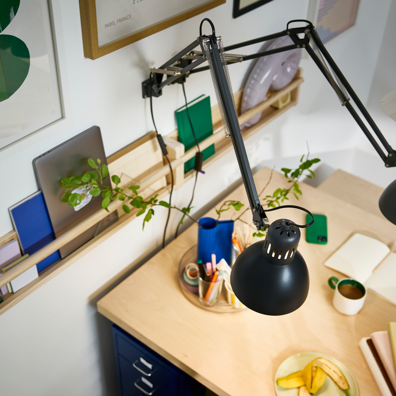 Two dark grey TERTIAL work lamps are attached to a wall with picture frames, shining their light over a BERGSHYTTAN table.