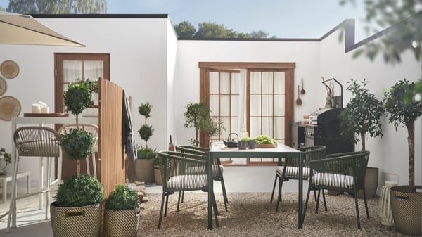 Leafy plants and SEGERÖN dark green/light grey table outdoor furniture on a bright, spacious, walled gravel-and-stone patio by a white, single-storey building. 