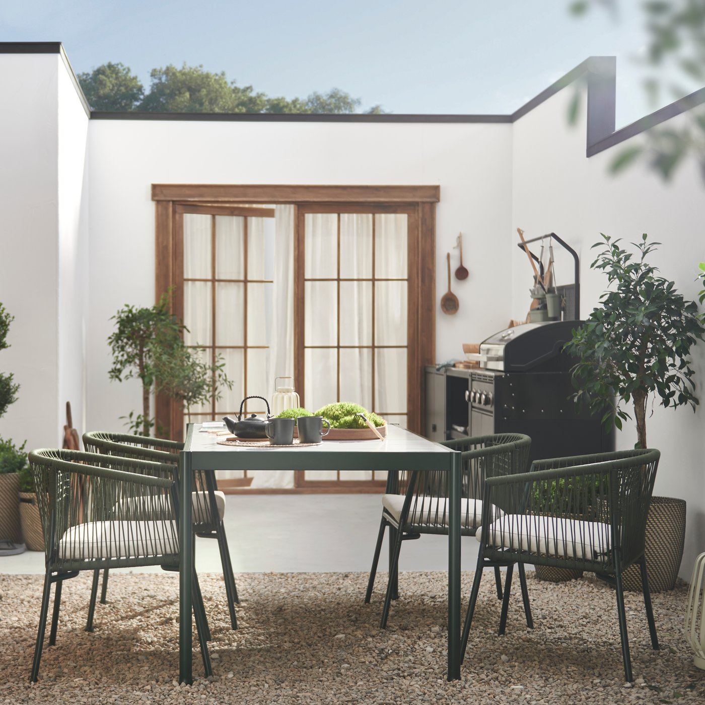 Leafy plants and SEGERÖN dark green/light grey table outdoor furniture on a bright, spacious, walled gravel-and-stone patio by a white, single-storey building. 