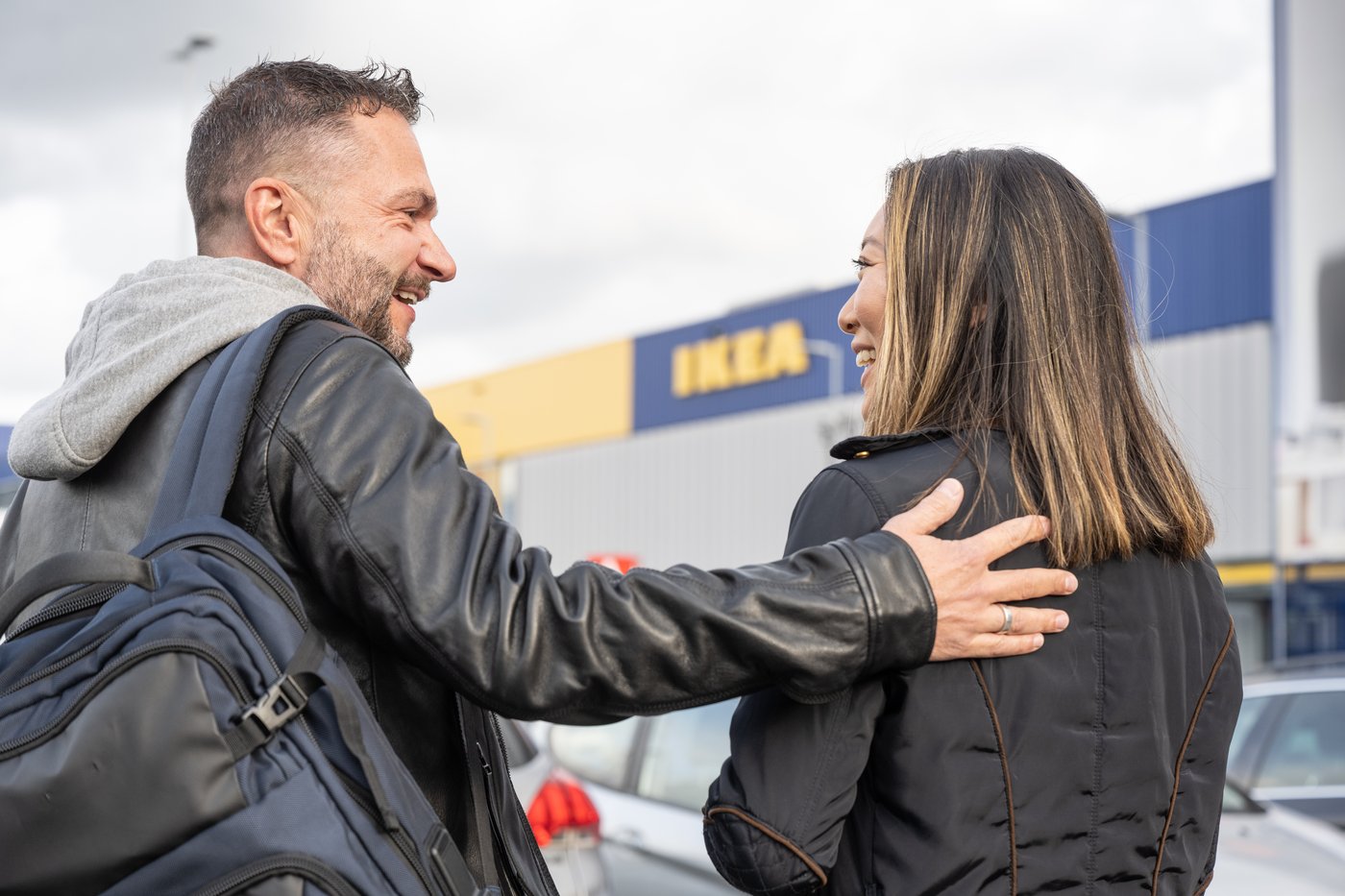 Två personer står utomhus framför en IKEA-butik. Personen till vänster har en ryggsäck och läderjacka och håller sin arm runt personen till höger, som har axellångt hår och är klädd i en mörk jacka. I bakgrunden syns bilar parkerade framför butiken.