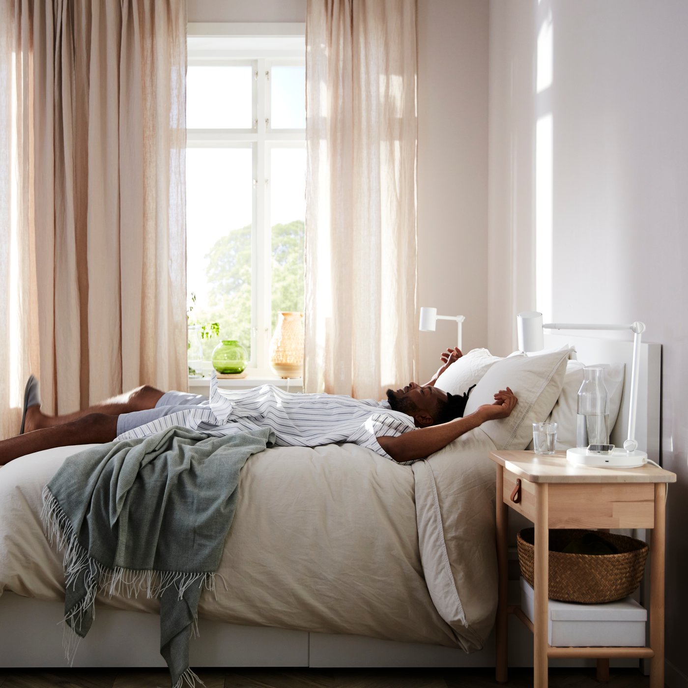 A person laying down on their back on a bed with beige bedding, in a sunny bedrooms with beige and sheer curtains.