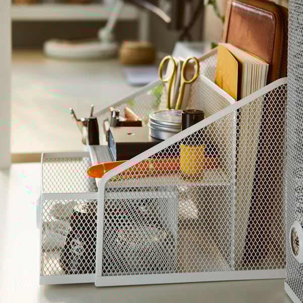 A DRÖNJÖNS desk organiser, holding many items such as a pen, a pair of scissors and a notebook, stands on a desk.