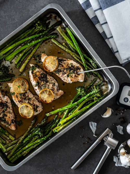 A rectangular, stainless steel KONCIS roasting tin with grill rack holds chicken breasts surrounded by asparagus.