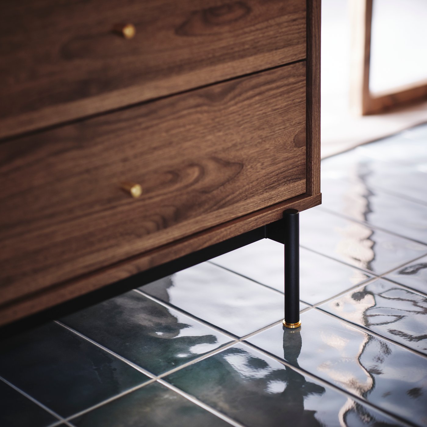 Close-up of RÅDMANSÖ chest of 5 drawers, with focus on brass knob and the black metal legs. Blue tile floor below.