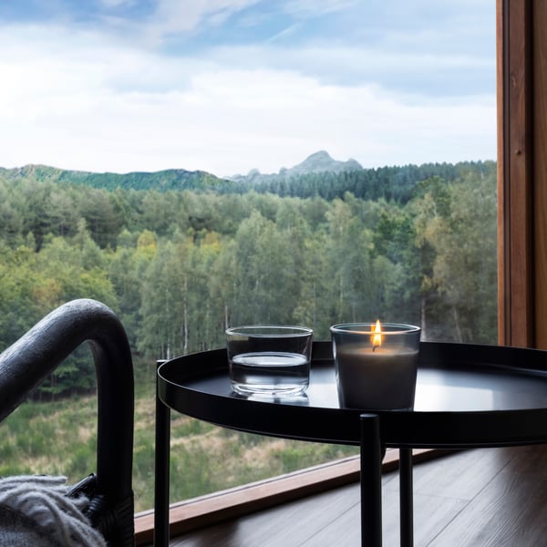 A PÄRONTRÄD scented candle with mountain scent stands on a black side table in front of a window. The view is a mountain with pine trees. 
