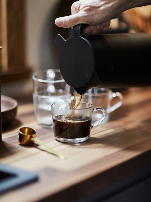 A hand pouring coffee from a thermos into an IKEA 365+ mug 24 cl in clear glass on a wooden table with other glass mugs.