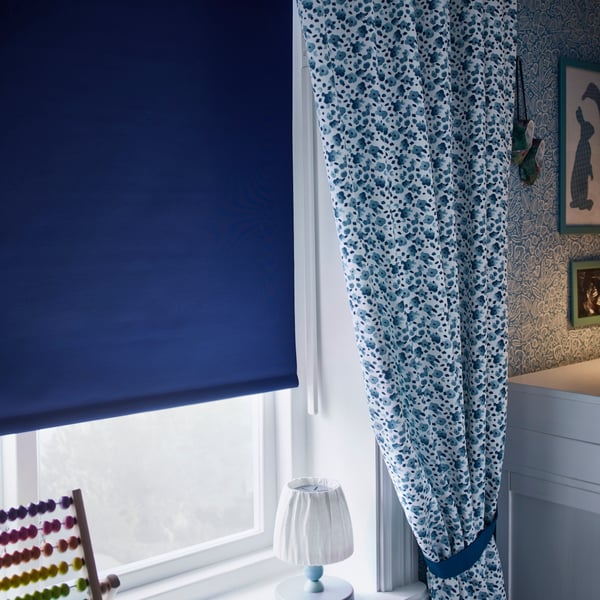 Cortina roller FRIDANS color azul, en una ventana con marco blanco. Además, hay una cortina de tela color blanco con diseños azules instalada por fuera de esta, amarrada a un lado de la ventana.