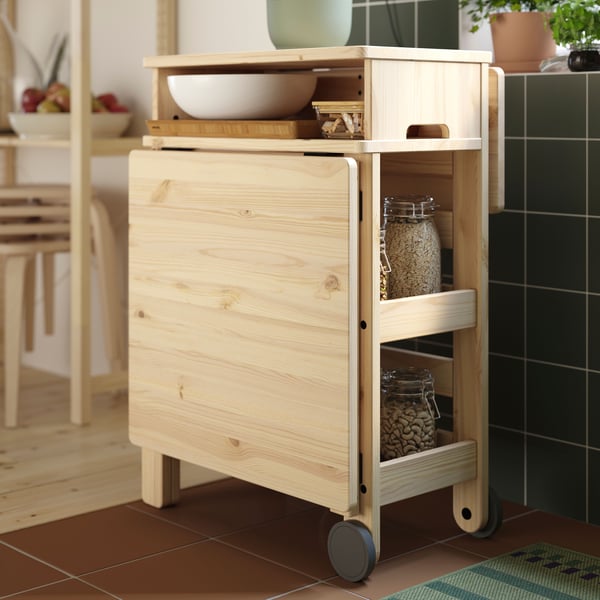 A kitchen has a RESARÖ mobile drop-leaf table with storage, folded down, with kitchenware stored on the open shelves.