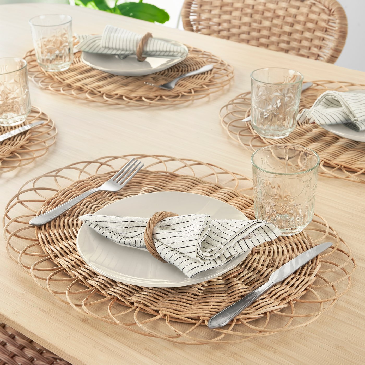Table set with woven placemats, white plates, striped napkins, glassware, and silverware on a light wood surface.