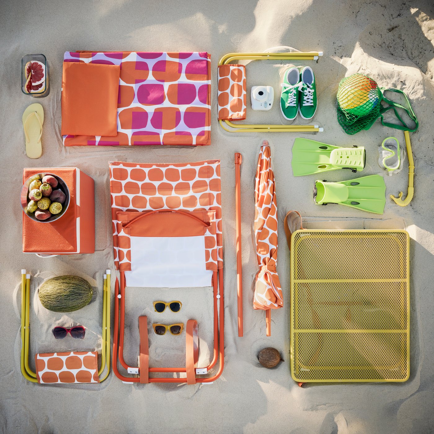 A beach setting with a bright orange STRANDÖN beach chair and various colourful accessories laid out on the sand.