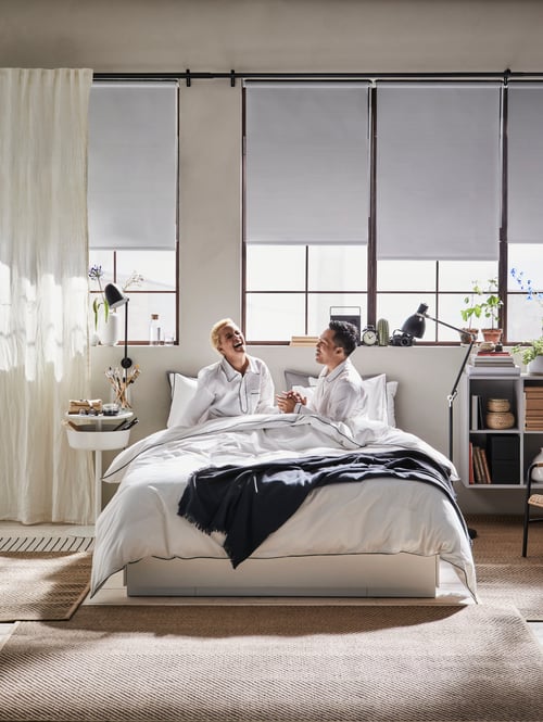 Ein Schlafzimmer mit einem NORDLI Bettgestell mit Schubladen und KUNGSBLOMMA Bettwäsche, einem EKET Schrank mit vier Fächern, einer ARÖD Stand-/Leseleuchte und FRIDANS Verdunklungsrollos.