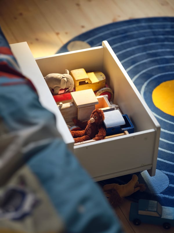A white SLÄKT storage box with castors filled with a mix of children’s toys, pulled out onto a round blue AFTONSPARV rug.