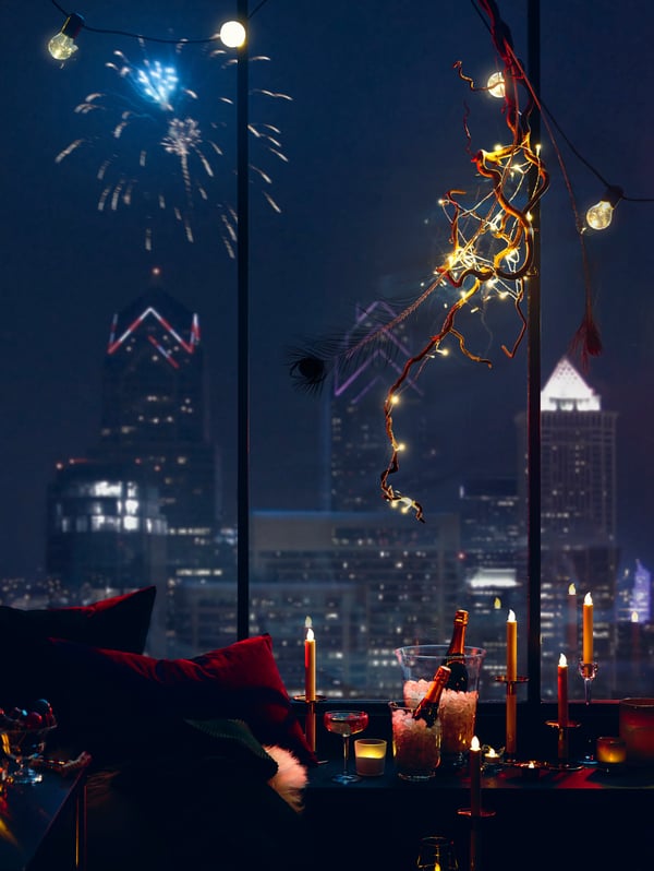 A window overlooking a city with fireworks in the background. There are fairy lights and candles in the apartment.