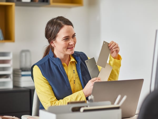 IKEA co-worker at a desk reviews material samples beside a laptop in an office setting, supporting planning and customer solutions.