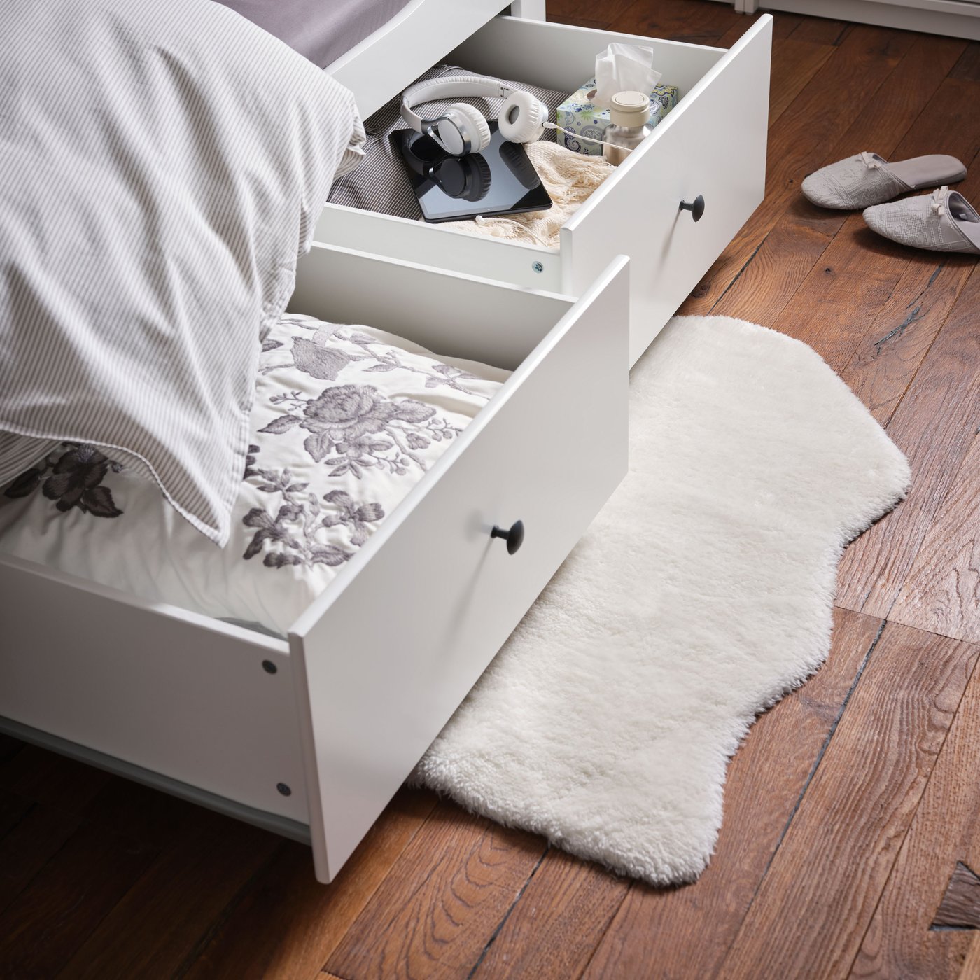 A white, sheepskin-style TOFTLUND rug made from recycled polyester is on the floor under a bed frame’s open drawers.