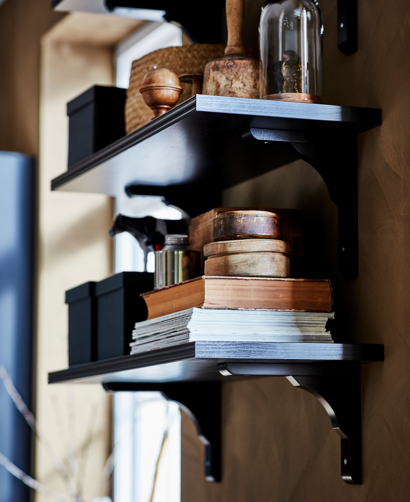 Zwei an die Wand montierte BERGSHULT/RAMSHULT Wandregale in Schwarz/Braun. Auf ihnen sind Bücher, Boxen und Dekogegenstände zu sehen.