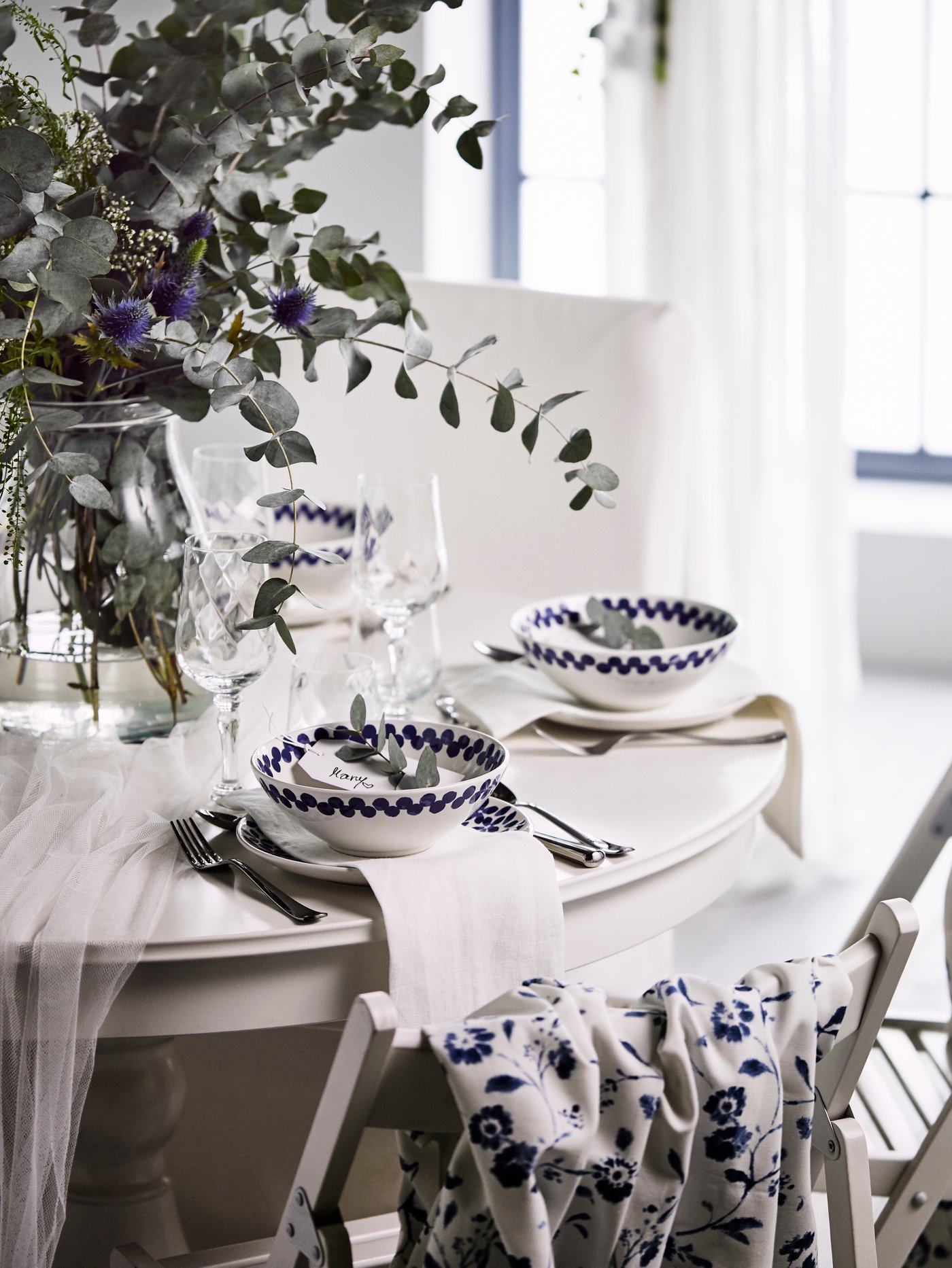 Ein weißer, festlicher runder Tisch mit Blättern und Wildblumen in einer Glasvase, Tellern und weiß-blauen MEDLEM-Schalen.