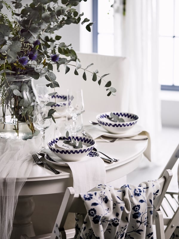 A white, festive round table with leaves and wild flowers in a glass vase, and plates and white and blue MEDLEM bowls.