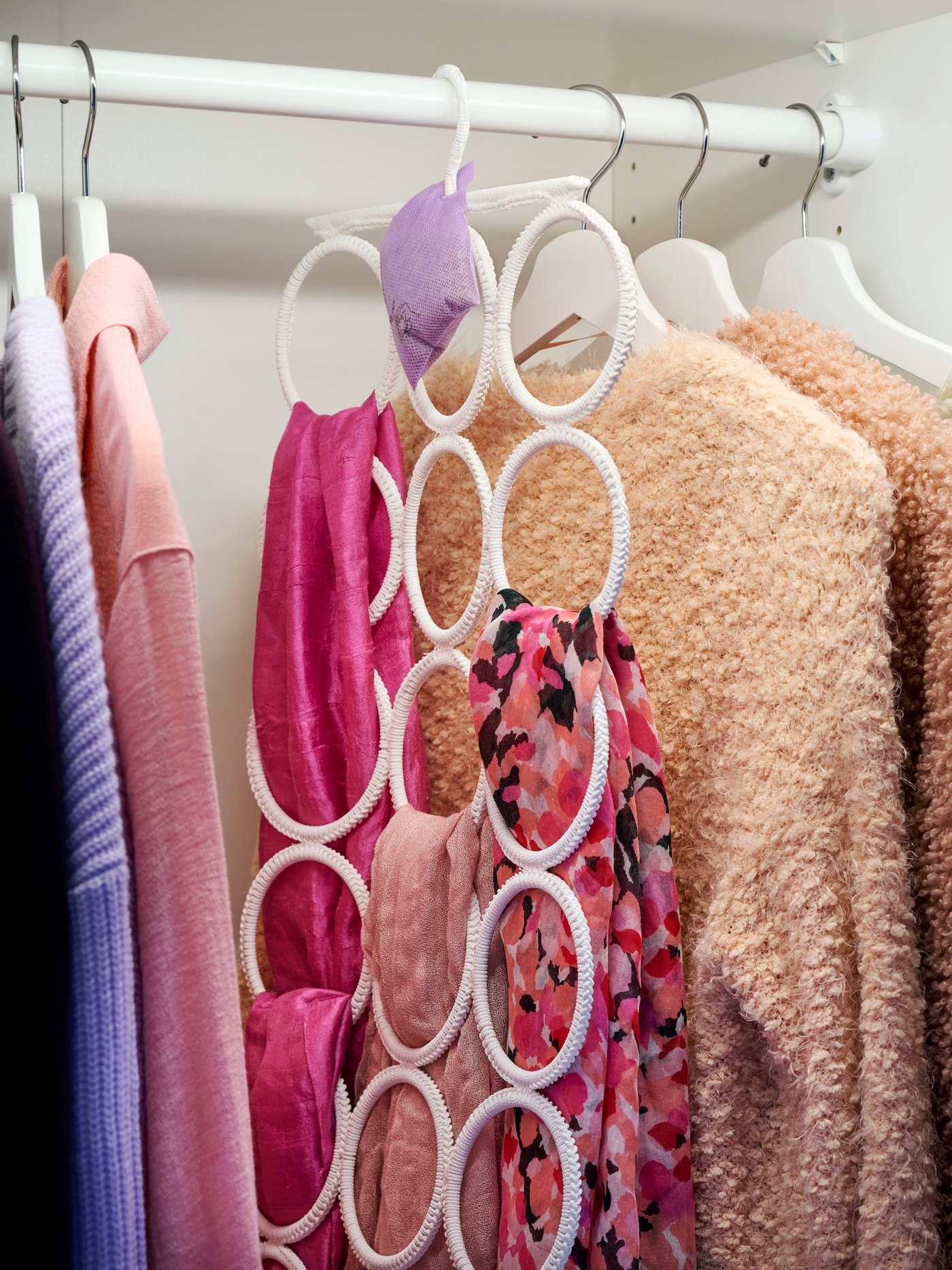 Primer plano del interior del armario con percha multiusos KOMPLEMENT con bufandas rosas y diferentes suéteres colgados.