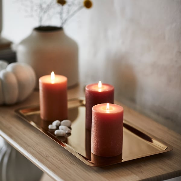 Tre orangea HÖSTAGILLE blockljus på en guldig bricka på ett sidobord.