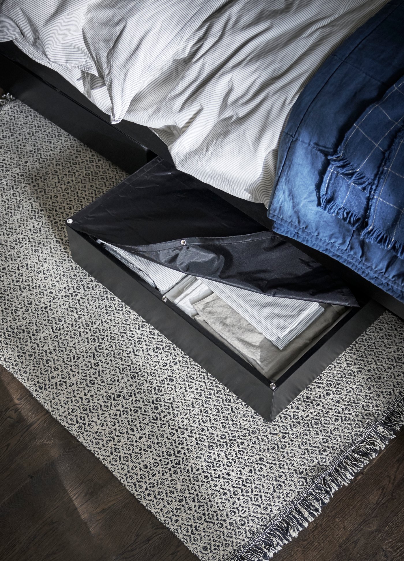 Een open lade onder een bed met witte lakens en een blauw deken toont netjes opgevouwen kleding, waaronder een wit hemd, naast een grijs vloerkleed met patroon.