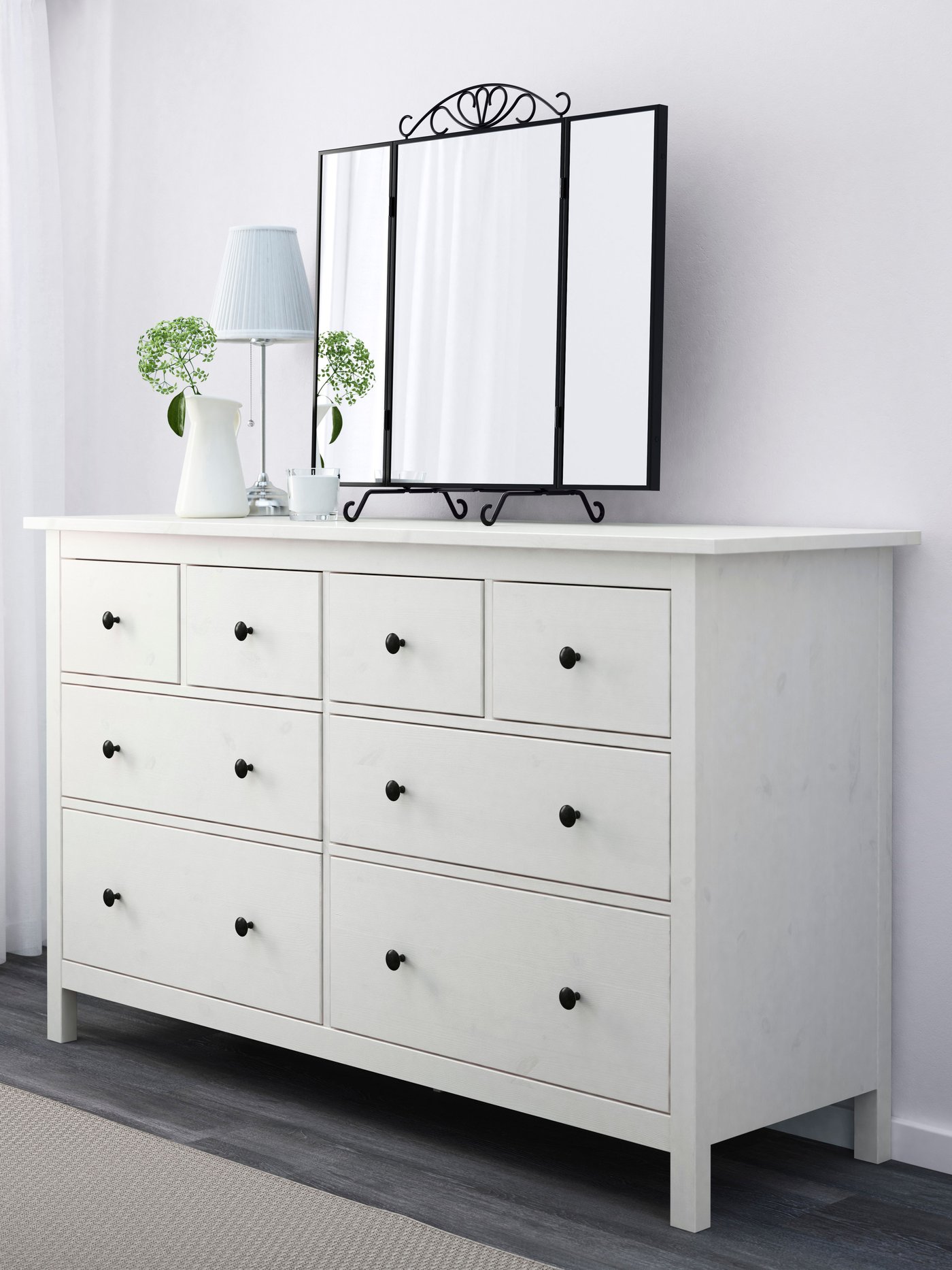 Op een zacht verlichte witte HEMNES ladekast met acht lades zien we een lamp, een plant en een spiegel.