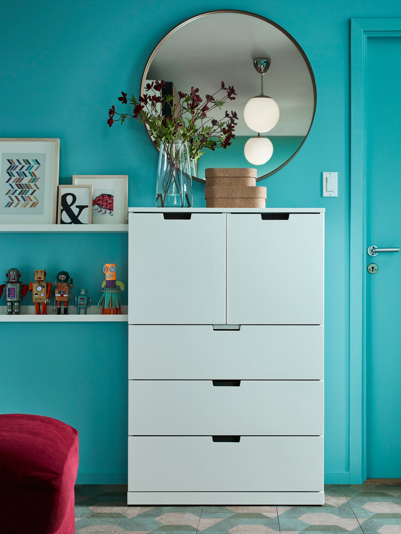 Una cómoda blanca NORDLI de cinco cajones con flores y cajas encima, junto a un espejo, una puerta y estantes abiertos con objetos decorativos.