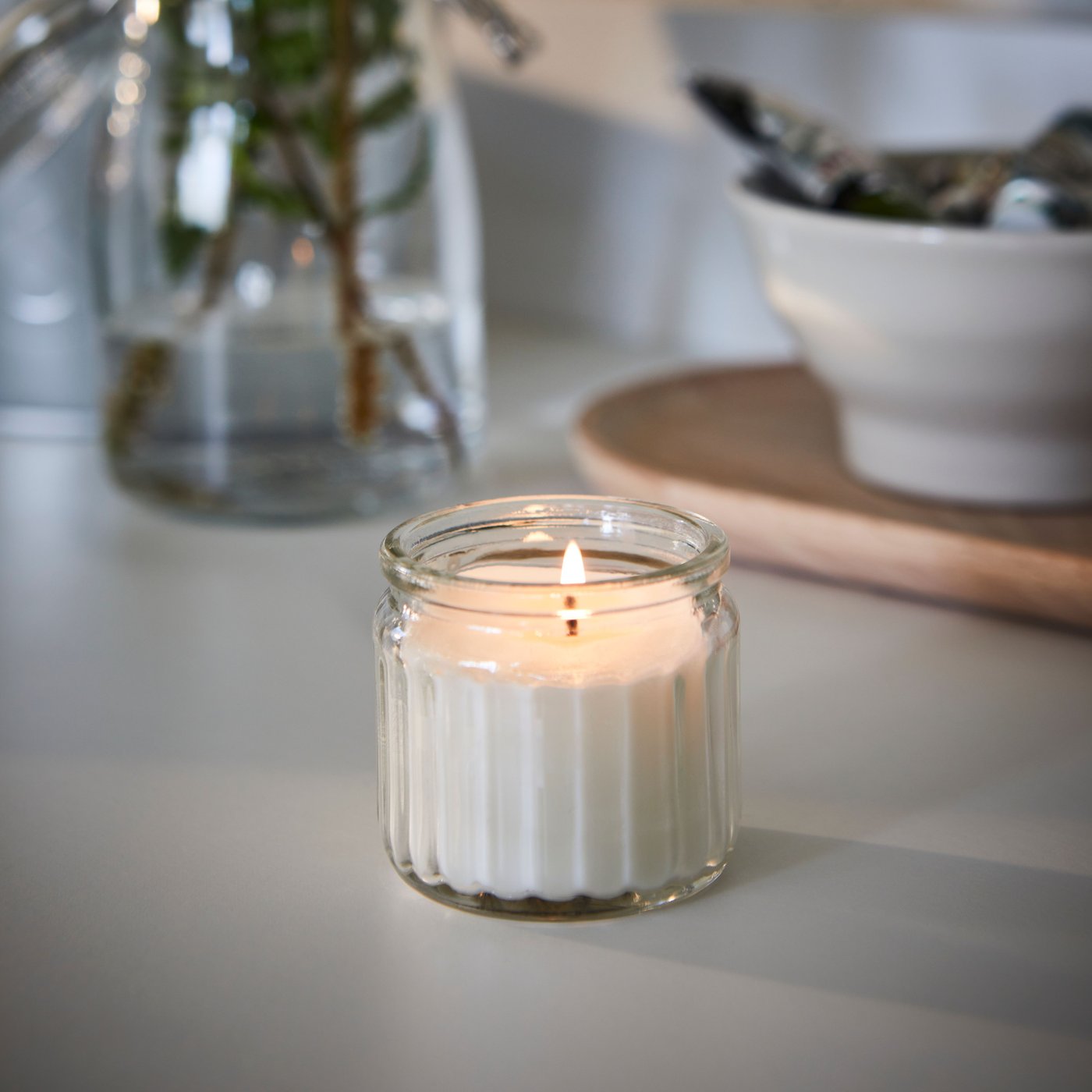 A Scandinavian Woods/white ADLAD scented candle in glass sits lit on a table.