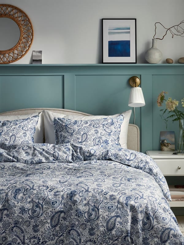 Pat cu lenjerie în nuanțe de alb și albastru, cu imprimeu floral, așezat pe un fundal de perete verde-albăstrui cu poliță decorată cu tablou, oglindă rotundă și vaze. Lampă de perete cu abajur alb, pentru seri liniștite în dormitor.