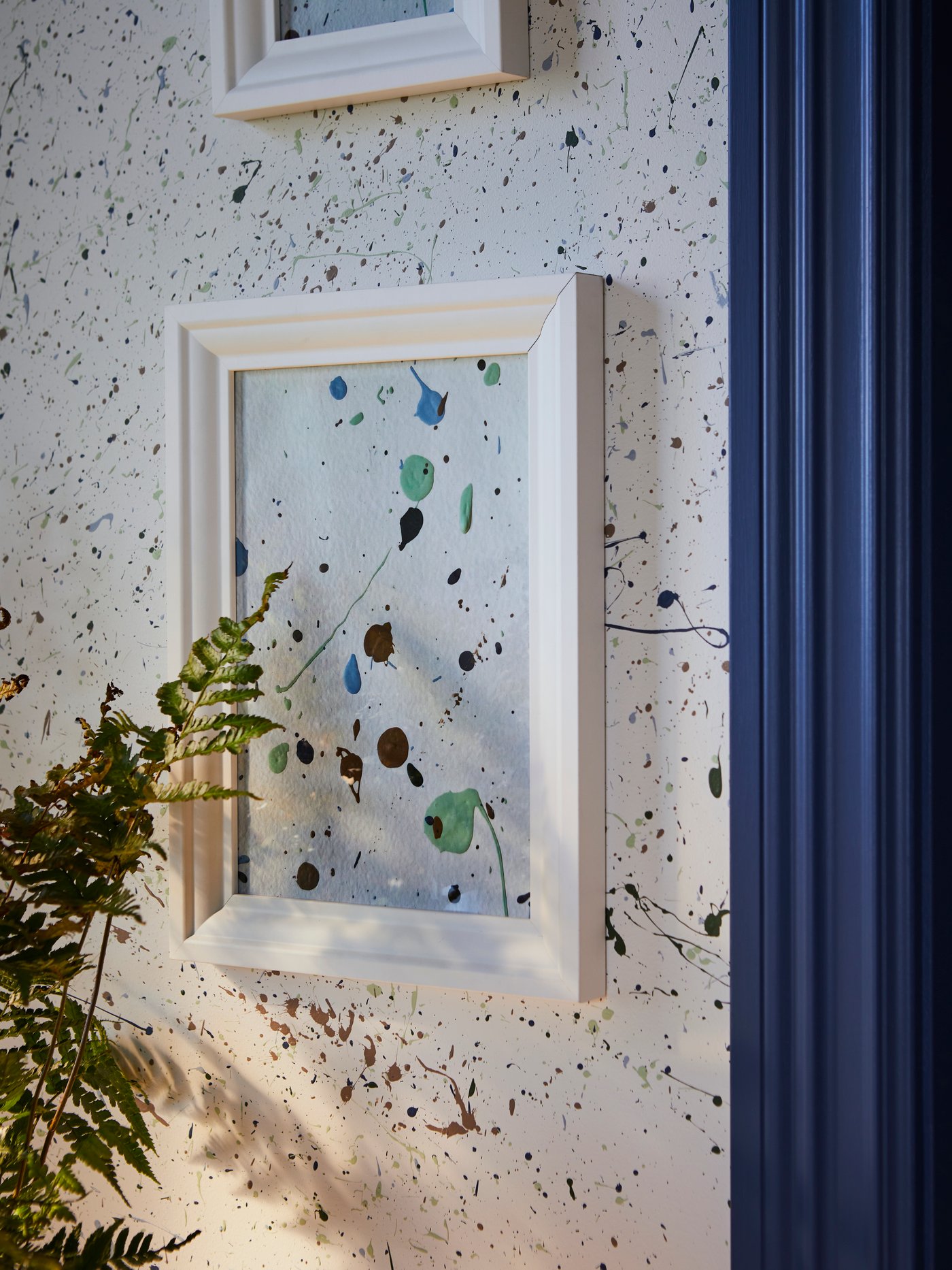 Dekorative Farbflecken in Kastanienbraun, Grün und Blau sind an einer weissen Wand und auf einem Stück Papier zu sehen, das in einem EDSBRUK Rahmen steckt.