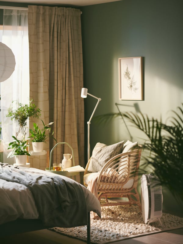 A BUSKO rattan armchair is in a corner of a green bedroom with plenty of plants, with a STARKVIND air purifier next to it.