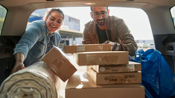 Een glimlachend stel laadt hun auto vol met IKEA-aankopen, waaronder verschillende kartonnen dozen en een groot opgerold tapijt, naast een herkenbare blauwe IKEA FRAKTA-tas.
