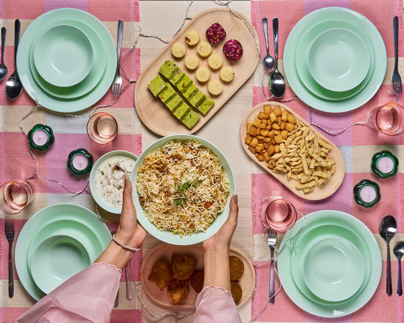 Overhead shot of a festive dining table with pastel green plates, rice dishes, sweets, and drinks on a pink tablecloth.