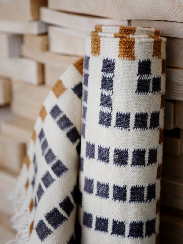 A VAMDRUP rug in shades of white, blue and brown, rolled up standing on its end leaning against a pile of timber.
