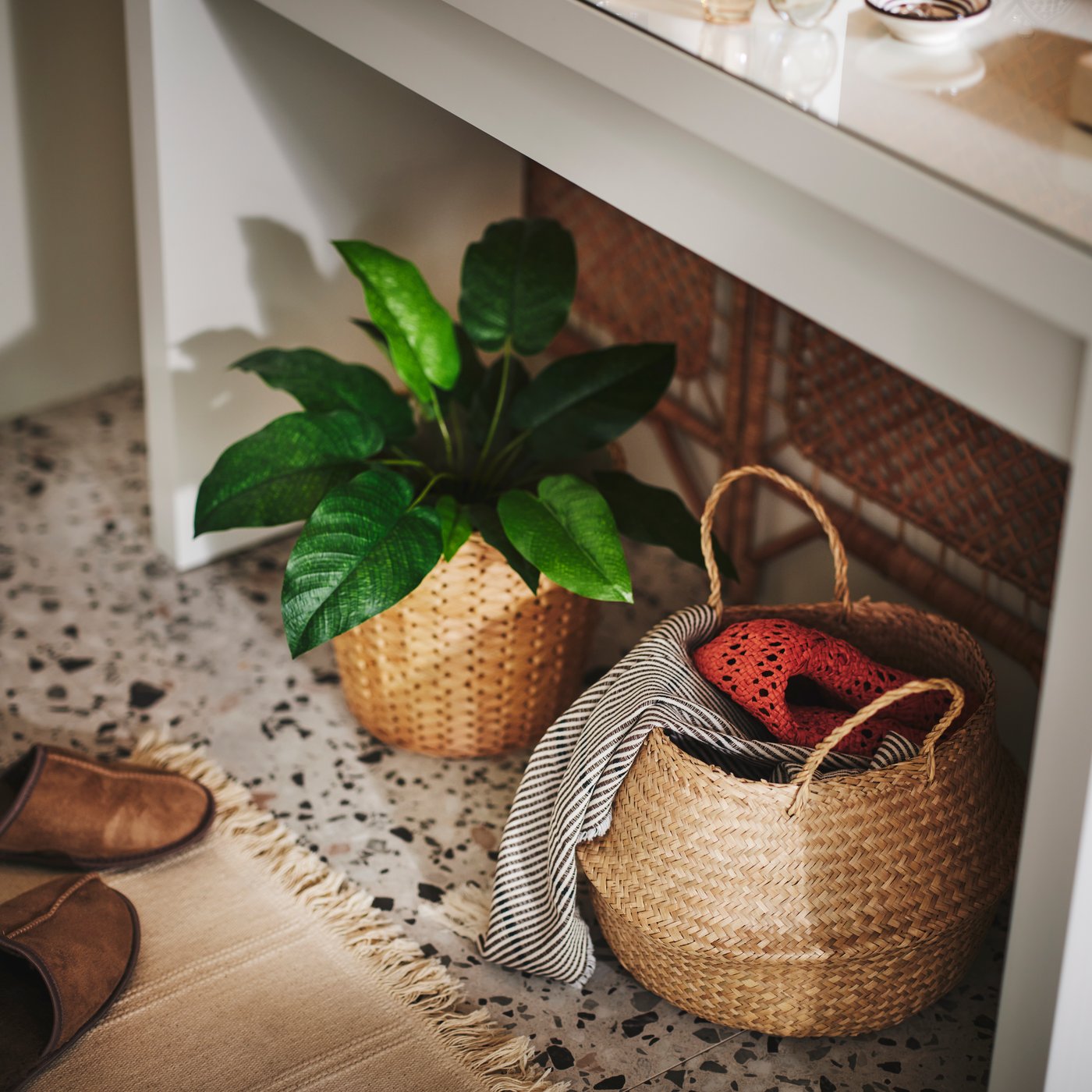 A FLÅDIS seagrass basket holding various textiles and a FRIDFULL plant pot on a rug on the floor under a dressing table.
