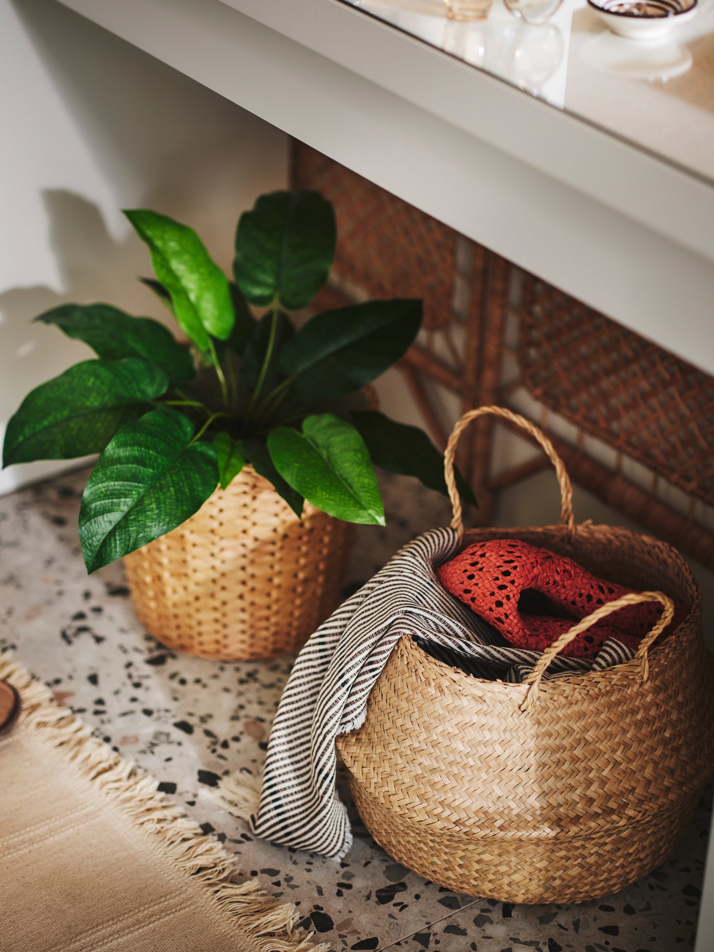 En FLÅDIS sjögräskorg som innehåller olika textilier och en FRIDFULL kruka på en matta på golvet under ett sminkbord.