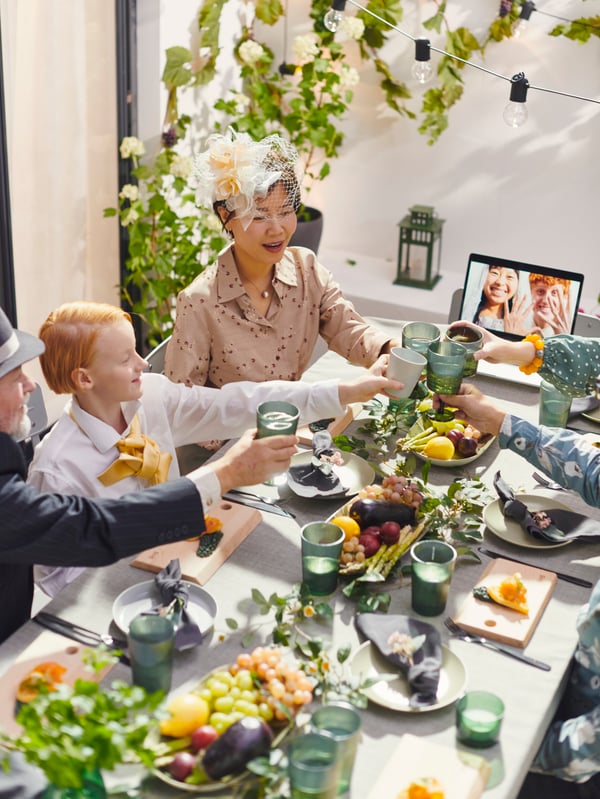 A large family clinks green glasses over a celebration table set up outside. A laptop with a newly married couple sits open.