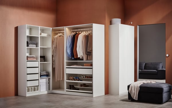 A white PAX wardrobe corner combination against a brown wall contains drawers, boxes, shoe racks and hanging clothes.