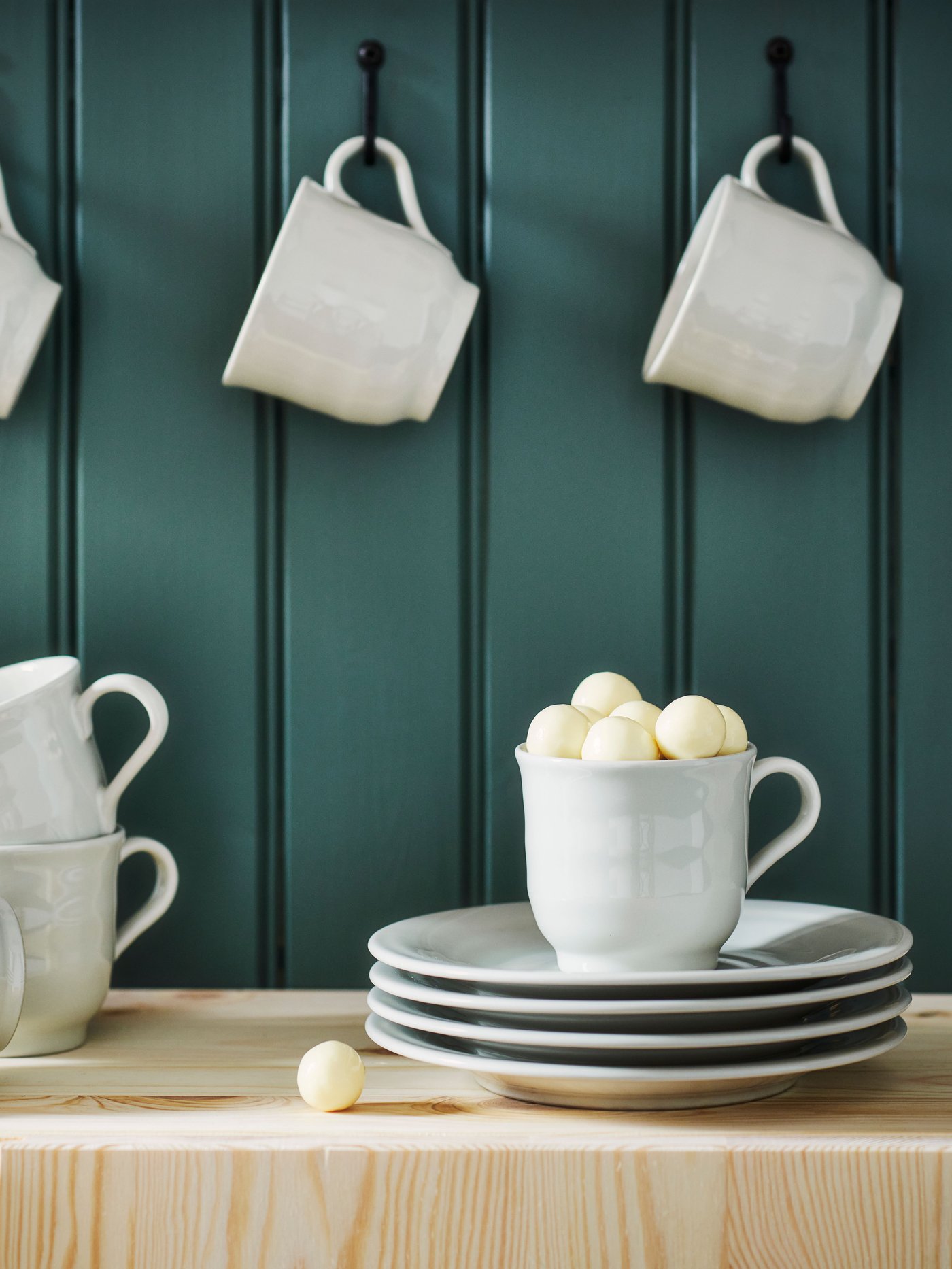 SANDSKÄDDA mugs hang from hooks on a wood panel wall. More mugs are underneath with a stack of SANDSKÄDDA side plates.